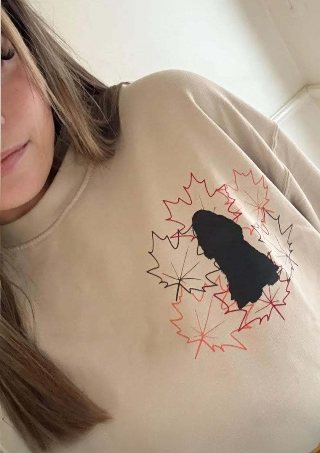 Person wearing a Beige Sweatshirt with a brown, orange and red maple leaf design with a dog in the middle.